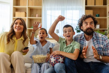 Neşeli aile evde spor maçı izlerken heyecanını paylaşıyor, keyifli oturma odasında birlikte tezahürat yapıyor ve mutluluğu ifade ediyor.