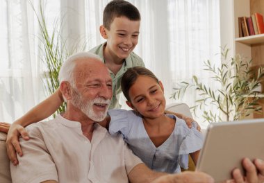 Büyükbaba, kız ve oğlanlarla vakit geçirmeyi seviyor. Tablete bakıp gülümsüyor. Parlak, davetkar bir oturma odasında.