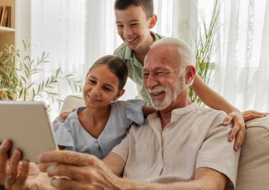Büyükbaba, kız ve oğlanlarla vakit geçirmeyi seviyor. Tablete bakıp gülümsüyor. Parlak, davetkar bir oturma odasında.