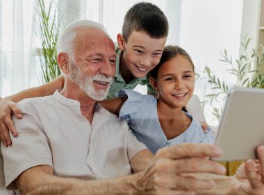 Büyükbaba, kız ve oğlanlarla vakit geçirmeyi seviyor. Tablete bakıp gülümsüyor. Parlak, davetkar bir oturma odasında.