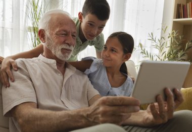 Büyükbaba, kız ve oğlanlarla vakit geçirmeyi seviyor. Tablete bakıp gülümsüyor. Parlak, davetkar bir oturma odasında.