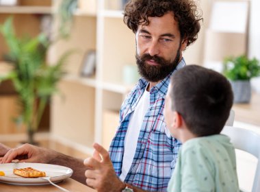 Baba ve küçük çocuk yemek masasında oturmuş, yemeğin tadını çıkarıyor ve sıcak yemek odasında sohbet ediyorlar.