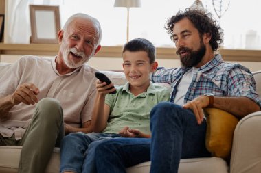 Üç nesil kanepede keyifli bir anı paylaşıyor, gülüyor ve ışık dolu sıcak oturma odasında televizyon seyrediyor.