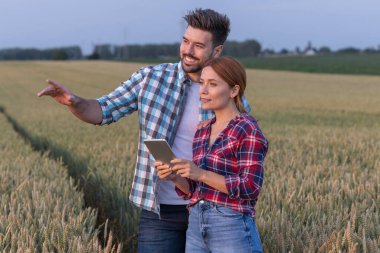 Çiftçi ve tarımcı buğday tarlasında dikilirken insan kameradan günbatımını gösteriyor. Kadının dikkatini tabletten uzaklaştırmak için elini omzuna koyar.