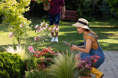 İnsanlar bahçede özenle çalışıyor. Çiçeklerin yanına çömelmiş ve mala kullanan kadına odaklı kesilmiş görüntü