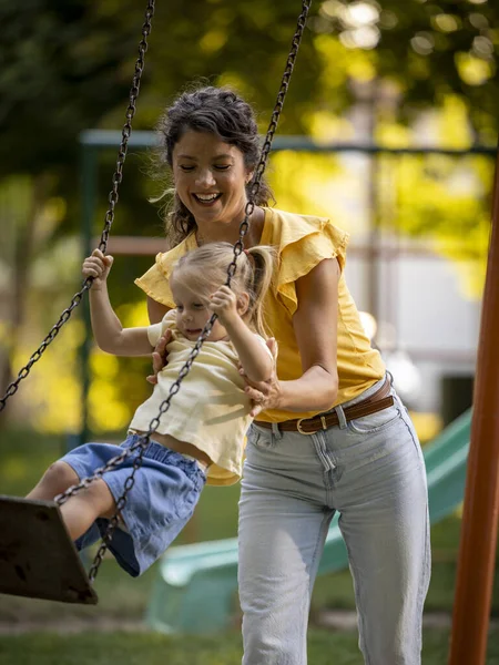Çocuk parkında sallanıyor. Neşeli genç anne kızının salıncakta durup eğlenmesine yardım ediyor. 