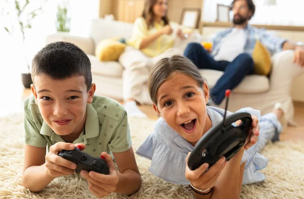 Yetişkinler kanepede dinlenirken iki çocuk halıda video oyunu oynuyor. Nişanlandılar ve eğleniyorlar.
