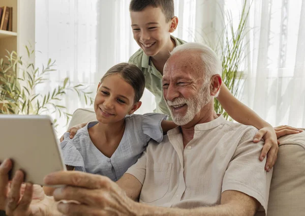 Büyükbaba, kız ve oğlanlarla vakit geçirmeyi seviyor. Tablete bakıp gülümsüyor. Parlak, davetkar bir oturma odasında.
