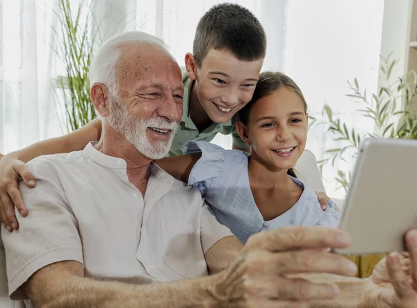 Büyükbaba, kız ve oğlanlarla vakit geçirmeyi seviyor. Tablete bakıp gülümsüyor. Parlak, davetkar bir oturma odasında.