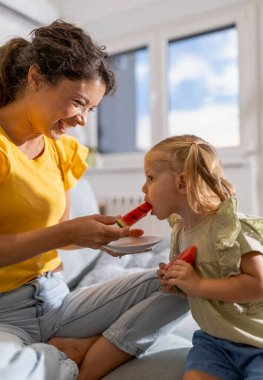 Neşeli genç kadın tatlı kızına oturma odasında bir dilim ferahlatıcı karpuz yediriyor.