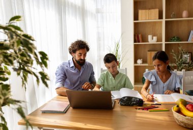 Baba, oğluna ve kızına ödevlerinde yardım ediyor yemek odasında dizüstü bilgisayarında kendi işini yapıyor. İyi ebeveynlik kavramı