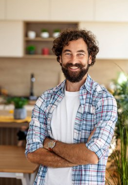 Kıvırcık saçlı ve sakallı bir adam parlak bir mutfakta duruyor, beyaz bir tişörtün üzerine kareli gömlek giyip güvenle gülümsüyor.