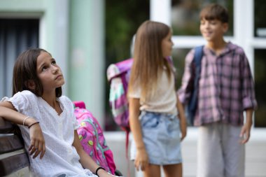 Okulun dışında üç genç öğrenci var. Bir kız bankta oturuyor ve kendini yalnız hissediyor. Arkasında iki öğrenci var, binanın yanında, konuşuyor ve gülümsüyor.