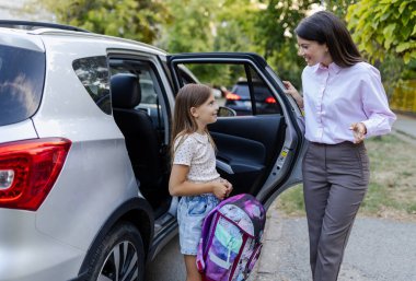 Sırt çantalı genç bir kız gümüş rengi arabadan çıkarken annesi açık arabanın kapısında durup gülümseyerek onunla okulun önünde konuşuyor.