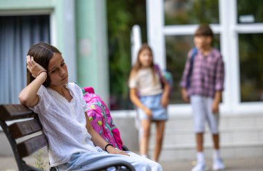 Okulun dışında üç genç öğrenci var. Bir kız bankta oturuyor ve üzgün hissediyor. Arkasında iki öğrenci var, binanın yanında, konuşuyor ve gülümsüyor.