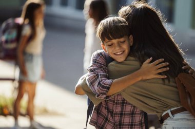 Genç çocuk okuldan sonra güneşli açık havada annesini kucaklarken diğer çocuklar da yakınlarda oyun oynuyor.