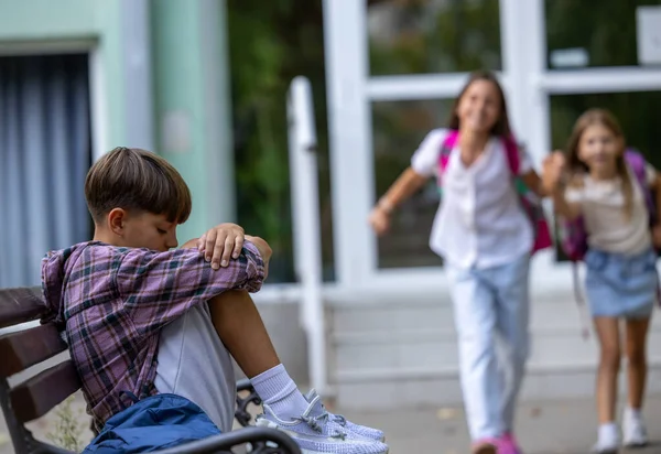 Güneşli bir okul günü açık hava ortamında iki kız arkasındayken oğlan bankta dalgın dalgın oturuyor.