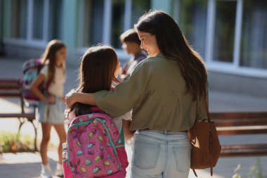 Anne arkası bakışı, okul dışında kızını kucaklıyor. Yakınlarda oynayan ve sohbet eden diğer çocuklar güneşli öğleden sonranın tadını çıkarıyorlar.