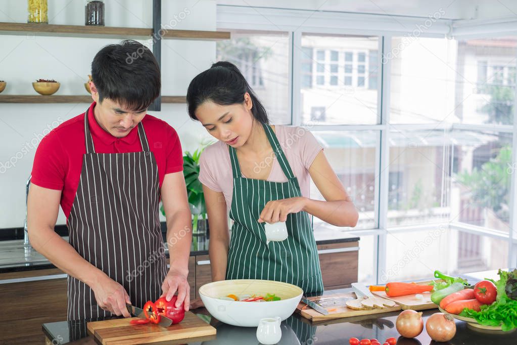 Asian couple's in apron, make salads together.  man are preparing to cut vegetables with knives. Woman mix salad dressing with vegetable  in big bowl, in kitchen. concept for foodie lover
