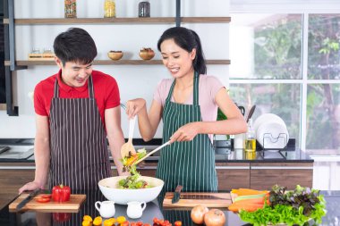 Asyalı genç çift önlük, birlikte yemek yapmak. Kadın büyük kase sebze ile salata sosu karıştırın. tezgahta, mutfakta, fotokopi alanında birçok bileşen vardır.
