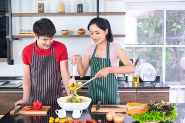 Asyalı genç çift önlük, birlikte yemek yapmak. Kadın büyük kase sebze ile salata sosu karıştırın. tezgahta, mutfakta, fotokopi alanında birçok bileşen vardır.
