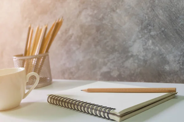 Focus of blank book, pencils white coffee cup on white desk.