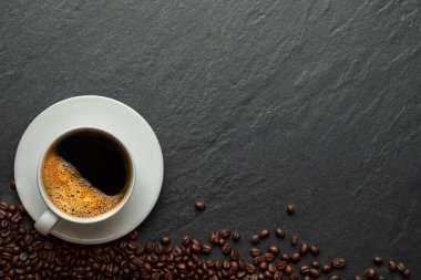 Süt köpüklü sıcak siyah kahvenin üstünde beyaz seramik fincanda kahve çekirdekleri kavrulmuş koyu taştan masa arkaplanında beyaz seramik bardakta. Metin için kopyalama alanı olan düz uzandı.