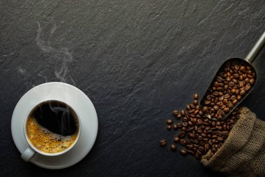 Siyah sıcak kahvenin üstünde duman ve beyaz seramik fincanda süt köpüğü ve kahve çekirdekleri koyu taş masa arkasında kavrulmuş. Kopyalama alanı olan düz.