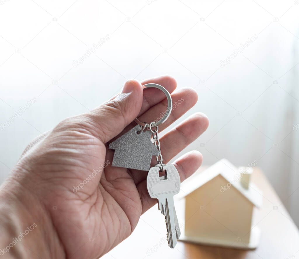 Close-up of hand holding silver keys house with house shaped keychain and home mockup on wooden table background. Real estate concept for buying a new home with copy space.