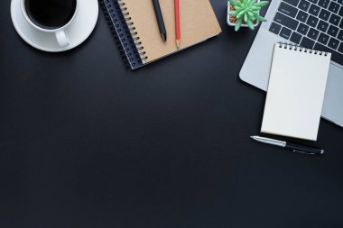Siyah ofis masasının üstünde dizüstü bilgisayar, defter ve kahve fincanı diğer ofis malzemeleriyle dolu. İş ve finans kavramı. Çalışma yeri, boş kopya alanı olan düz uzanma.