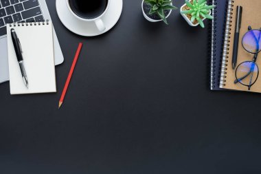 Siyah ofis masasının üstünde dizüstü bilgisayar, defter ve kahve fincanı diğer ofis malzemeleriyle dolu. İş ve finans kavramı. Çalışma yeri, boş kopya alanı olan düz uzanma.