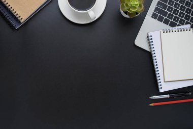 Siyah ofis masasının üstünde dizüstü bilgisayarı, not defteri ve ekipman ofis malzemeleriyle dolu kahve fincanı var. İş ve finans kavramı. Çalışma yeri, boş kopya alanı olan düz uzanma.