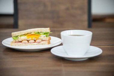 Sandviç ve beyaz tabak ve ahşap masa üzerinde kahve fincan, sandviç ekmek, sosis, kızarmış yumurta ve vetgetable yapılır Amerikan kahvaltı veya fast food sağlıksız gıda veya abur cubur olduğunu