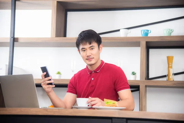 O homem de negócios com casual t-shirt vermelha mensagens no telefone móvel, beber café e comer sanduíche, Jovem trabalhando em labtop ou computador na sala de cozinha, estilo loft — Fotografia de Stock