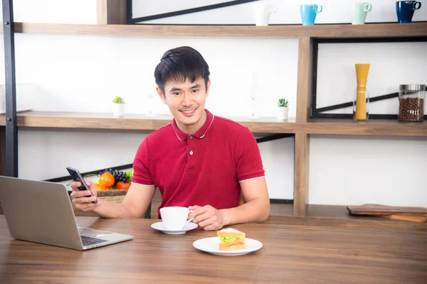 O homem de negócios com casual t-shirt vermelha mensagens no telefone móvel, beber café e comer sanduíche, Jovem trabalhando em labtop ou computador na sala de cozinha, estilo loft — Fotografia de Stock