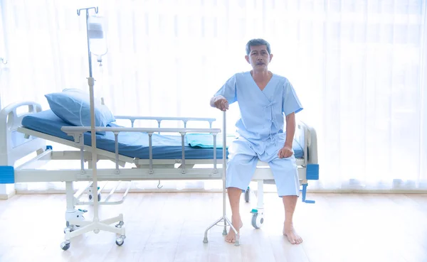 Asian sick  or elderly  old man couldn't walk sitting alone on the patient's bed with blue shirt and walking stick waiting for the doctor and nurse for treatment or cure in the hospital or healthcare