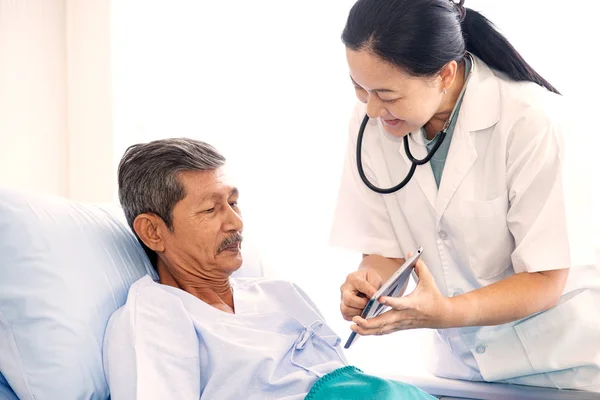 Азиатская женщина профессиональный врач с планшетом посещения и диагностики старика пациента, лежащего в постели в больничном отделении. Врач имеет рутинную работу, чтобы проверить и консультировать пациента — стоковое фото