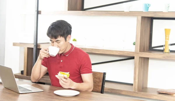 Asiático sorrindo jovem com casual camiseta vermelha desfrutar de tomar café da manhã, comer sanduíche e trabalhar com notebook, Jovem cozinhando comida e bebida na sala de cozinha estilo loft — Fotografia de Stock