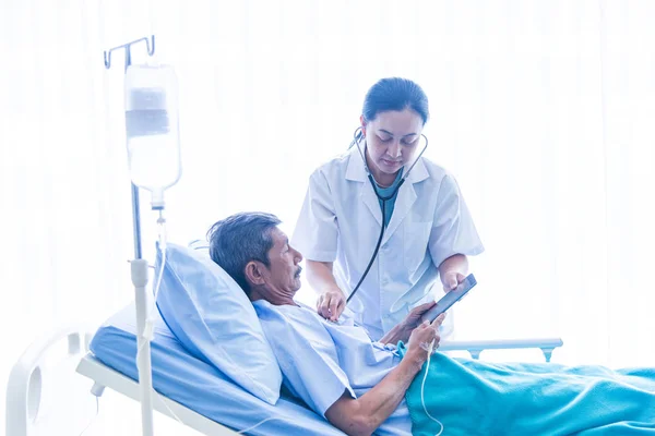 Азиатская женщина-врач с блокнотом улыбается, консультирует, говорит, и диагностики пациента старика, лежащего в постели пациентов в больнице палаты — стоковое фото