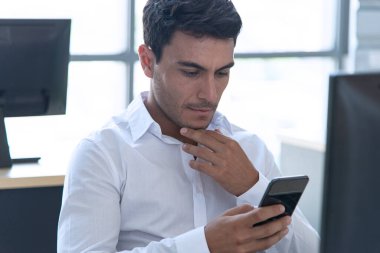 Kendine güvenen genç yönetici, ciddi olarak bilgisayar üzerinde çalışan ve modern ve yaratıcı ofiste akıllı telefon kullanan bir yönetici. Sosyal uzaklık durumunda çalışmak.