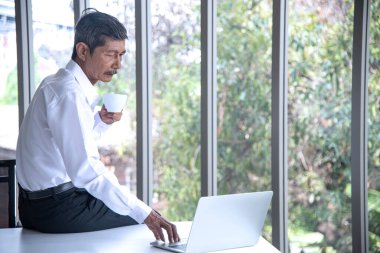 Evden ve sosyal uzaklık kavramından çalışmak. Asyalı iş adamı çalışma alanında çalışıyor ve kahve içiyor. Yaşlanan adam çevrimiçi dizüstü bilgisayar toplantısı kullanıyor.