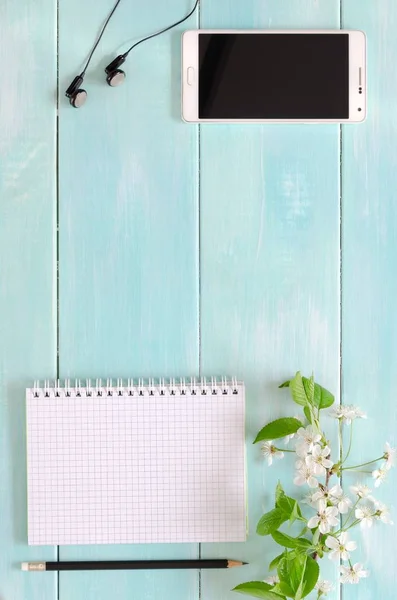 Open notebook with pencil, phone, headphones and sprig with cherry blossoms on mint wooden background. Copy space background, top view flat lay overhead