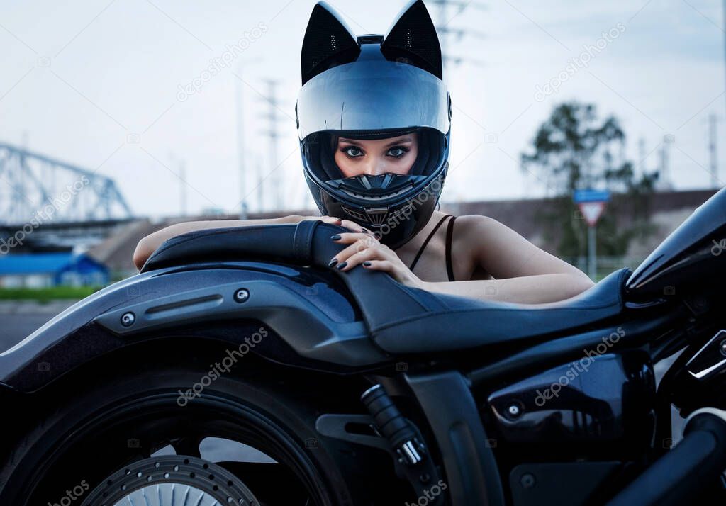 A beautiful girl in a helmet looks from behind a motorcycle.