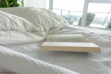 Uzun pencereler ve pencere ışığı ile parlak bir daire yatak odasında kırışık, kırmızı duvar sanatı ile kullanılan yatak kitap. Evde veya hafta sonu dinlenmek, yatakta kitap okumak.