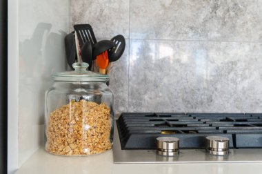 Jar of homemade granola next to modern gas stovetop with kitchen utensils and marble tile backsplash in the background. Bright, daylight. Breakfast food. clipart