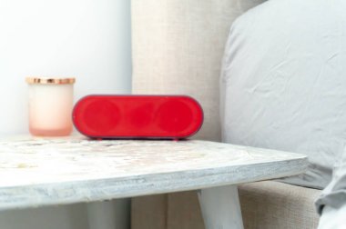 Parlak kırmızı radyo çalar saat ve mum kavanozu bir başucu komidi üzerinde, yatak ve çarşafyumuşak renklerde gösteren.