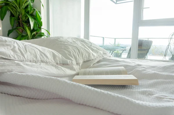 Reading in bed, staying in bed. Wrinkled sheets in a used bed, in a real home bedroom. Apartment windows overlooking a balcony, with bright light entering the room. White linens. — Stock Photo, Image