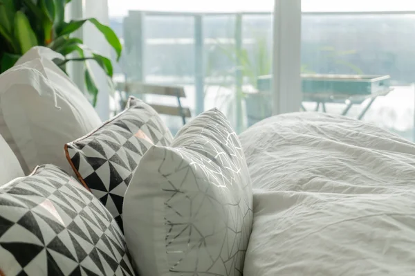 Fluffy, decorative pillows on a real bed, with wrinkled sheets and grey, black and white accent colors. Balcony view shows in the background, as well as a house plant.