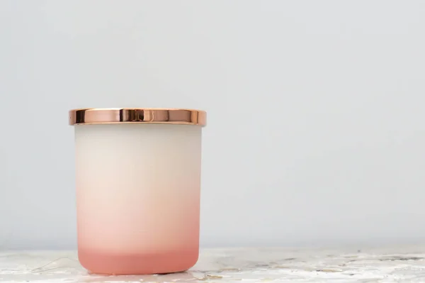 Rose pink and white ombre candle jar with a shiny gold lid, sitting on a bedside table (night stand). Candle is not lit. Daytime.