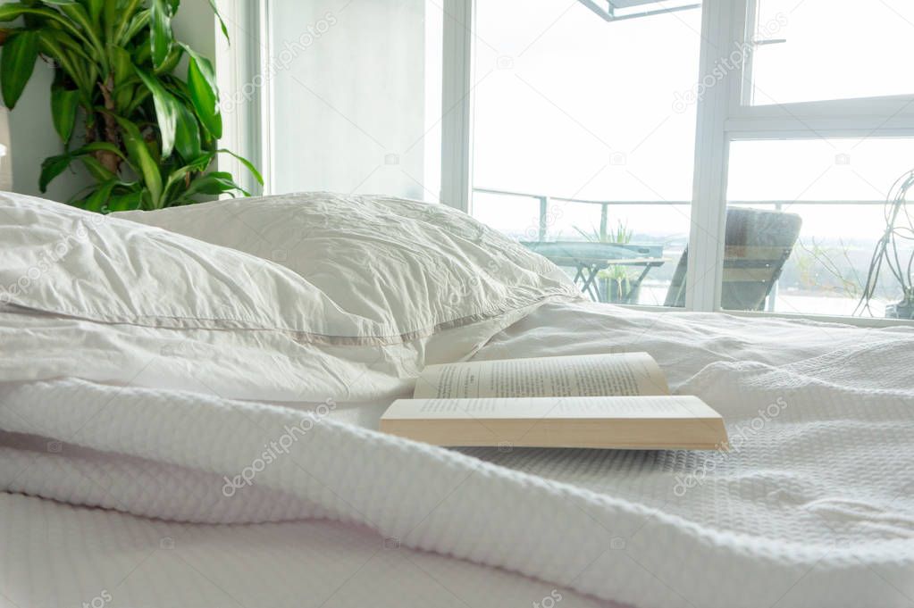 Reading in bed, staying in bed. Wrinkled sheets in a used bed, in a real home bedroom. Apartment windows overlooking a balcony, with bright light entering the room. White linens.